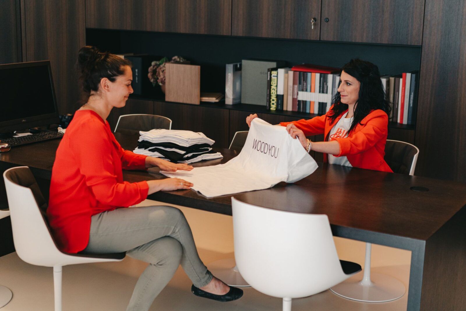 Twee vrouwen bespreken logo op kleding in een kantoor in Houthalen, met textielbedrukking in Limburg en promotieartikelen op tafel.