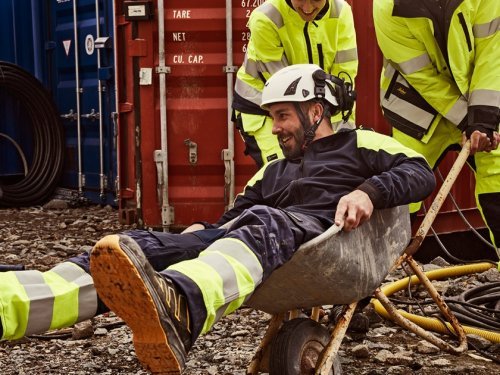 Bouwvakker geniet van een pauze in een kruiwagen, omringd door felgele signalisatiekleding. Val op met onze signalisatiekleding!