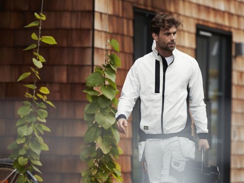 Man in witte jas loopt langs een modern gebouw met groene planten, passend bij de slogan "Voor ieder wat wits!" voor veelzijdige en stijlvolle mode.