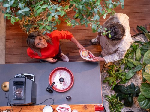 Twee mensen in gesprek bij een keuken, omringd door planten. Samenwerking met Limburgs Landschap: Bedrijfskleding met een Verhaal.